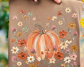 May include: A brown t-shirt featuring a central orange pumpkin illustration surrounded by a variety of painted-style flowers in shades of pink, orange, yellow, and white. The design uses thick, textured brushstrokes to create a dimensional effect.