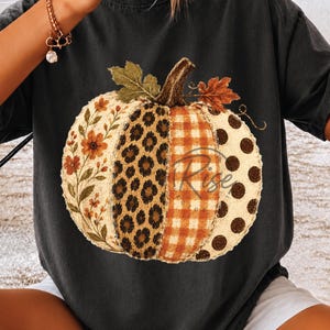 May include: A charcoal gray t-shirt featuring a large pumpkin graphic. The pumpkin is divided into four vertical sections with floral, leopard print, orange gingham, and brown polka dot patterns, topped with green and brown leaves.