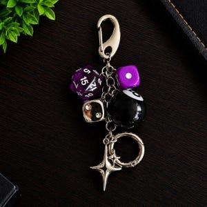 May include: A silver-toned metal keyring featuring a purple translucent twenty-sided die, a solid purple cube die, a black eight ball, a silver metallic cube die with clear stones, and a four-pointed star charm attached to a lobster claw clasp.