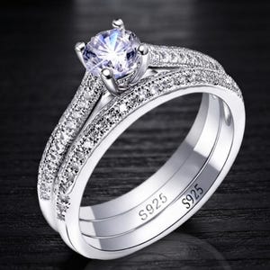 May include: A silver-toned ring set featuring a central round-cut clear stone held in a four-prong setting. The band and the accompanying ring are encrusted with small clear stones along the top surface. The inner band is engraved with S925.