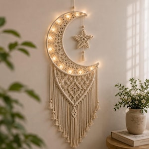 May include: A cream-colored macrame wall hanging shaped like a crescent moon with an attached star. The moon frame is woven with intricate cord patterns and illuminated by small warm white LED lights. Long fringe tassels hang from the bottom.