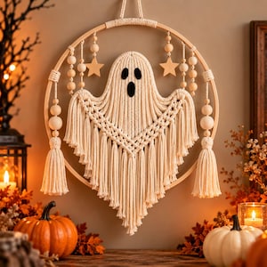 Puede incluir: Un tapiz de macramé con una figura de fantasma central hecha de cuerda de algodón color crema, suspendida dentro de un aro de madera circular. El diseño incluye cuentas de madera y dos pequeñas estrellas de madera, con flecos largos.