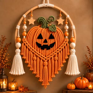 May include: A macrame wall hanging featuring an orange pumpkin with a black jack-o'-lantern face, suspended from a wooden hoop. The design includes green cord leaves, cream-colored tassels, wooden beads, and two wooden star accents.
