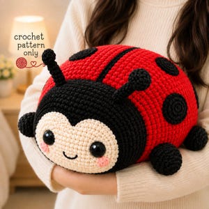 Puede incluir: Un juguete de peluche de mariquita hecho a ganchillo con el dorso rojo brillante y puntos negros. La cabeza es negra con la cara color crema, dos antenas negras y dos ojos negros. El texto "crochet pattern only" aparece en la esquina superior izquierda.