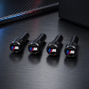 May include: Four black cylindrical valve stem caps featuring the M logo in white with blue and red stripes on the circular top. Each cap has a smooth, matte finish and a grooved design along the side, presented in a set of four identical pieces.