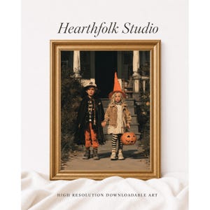Peut inclure: Une impression encadrée intitulée Hearthfolk Studio montrant deux enfants en costumes d'Halloween sur un porche. L'un porte une cape noire et un pantalon orange, l'autre une veste beige, des leggings rayés et un chapeau de sorcière orange, tenant une citrouille.