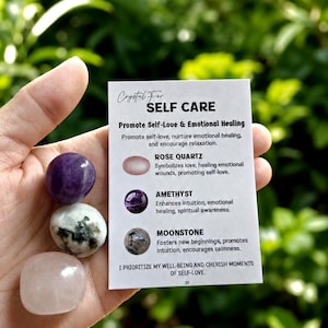 May include: Three polished tumbled stones including a deep purple amethyst, a white and grey speckled moonstone, and a pale pink rose quartz, held next to a white informational card titled Crystal For SELF CARE with text descriptions.