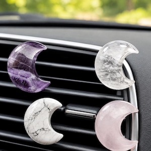 May include: Four crescent moon-shaped stone ornaments attached to a black car air vent. The set includes a translucent purple amethyst, a clear quartz, a white stone with black veining, and a smooth, solid light pink rose quartz piece.
