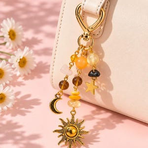 May include: A gold-toned metal keyring featuring a lobster clasp, three dangling chains with assorted beads in shades of yellow, orange, brown, and clear, and charms shaped like a crescent moon, a five-pointed star, and a detailed sun.