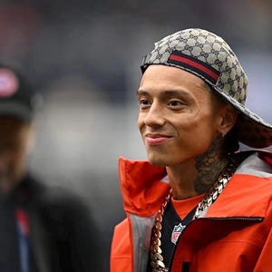 May include: A beige baseball cap with a repeating dark brown logo pattern and a horizontal red and green stripe across the front, worn with a bright orange jacket and a thick silver chain necklace featuring a prominent metallic link design.