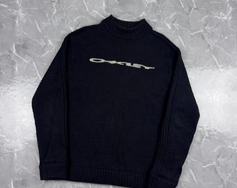 May include: A black long-sleeve sweater with a ribbed mock neck collar, cuffs, and hem. The front of the sweater features the word Oakley embroidered in a light-colored, textured font across the chest area, with vertical ribbed detailing on the sleeves.