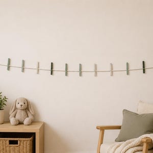 May include: A wall-mounted display garland featuring a long cord strung with multiple small, vertical wooden clips in alternating shades of sage green, dark forest green, and cream, finished with circular wooden beads at each end.