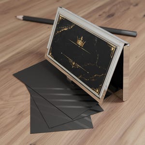 May include: A silver-toned metal business card holder featuring a black marble-patterned insert with gold-colored accents, including a crown graphic and decorative lines. The holder is displayed with several plain black cards underneath it.