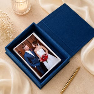 May include: A deep blue fabric-covered box containing a stack of rectangular photographs. The top photo shows a person in a navy blue suit and a person in a white strapless wedding dress holding a red floral bouquet. A gold pen lies nearby.