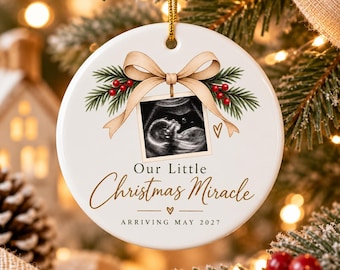 Könnte beinhalten: Ein rundes, weißes Keramikornament mit einem zentralen Ultraschallbild, das an einer beigen Schleife mit grünen Tannenzweigen und roten Beeren hängt. Darunter steht der Text Our Little Christmas Miracle und ARRIVING MAY 2027.