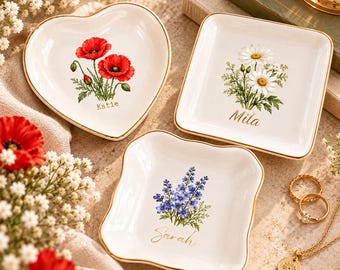 Könnte beinhalten: Drei weiße Keramikschalen für Schmuck mit goldfarbenen Rändern. Eine herzförmige Schale zeigt rote Mohnblumen und den Namen Katie. Eine quadratische Schale zeigt weiße Gänseblümchen und den Namen Mila. Eine quadratische Schale zeigt blaue Blumen und Sarah.