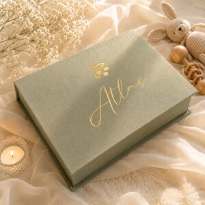 May include: A rectangular sage green keepsake box with a hinged lid featuring a gold foil mobile icon and the name Atlas in cursive script. The box is positioned on a soft fabric surface alongside a small crocheted rabbit and a wooden rattle.