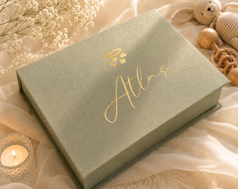 May include: A rectangular sage green keepsake box with a hinged lid featuring a gold foil mobile icon and the name Atlas in cursive script. The box is positioned on a soft fabric surface alongside a small crocheted rabbit and a wooden rattle.