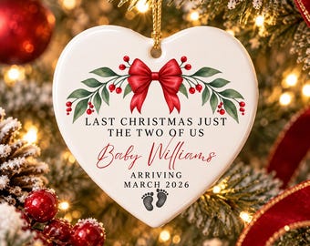 Könnte beinhalten: Ein weißes, herzförmiges Keramikornament mit einer roten Schleife, grünen Stechpalmenblättern und roten Beeren. Der Text lautet: LAST CHRISTMAS JUST THE TWO OF US, Baby Williams, ARRIVING MARCH 2026, mit zwei kleinen schwarzen Babyfußabdrücken.