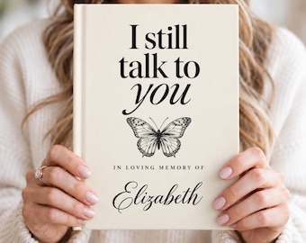 Könnte beinhalten: Ein cremefarbenes Hardcover-Buch mit dem schwarzen Text I still talk to you, einer detaillierten schwarzen Schmetterlings-Strichzeichnung, dem Text IN LOVING MEMORY OF und dem Namen Elizabeth in Schreibschrift.