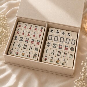 May include: A cream-colored storage box containing a set of off-white mahjong tiles. The tiles feature intricate black, red, and green engraved characters, numbers, and floral illustrations. The set is organized into two compartments within the box.