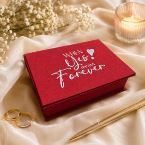 May include: A rectangular red box with a textured finish featuring the silver text "WHEN Yes! means Forever" and a small silver heart icon. The box is positioned on a draped cream fabric alongside two gold rings and a gold ballpoint pen.