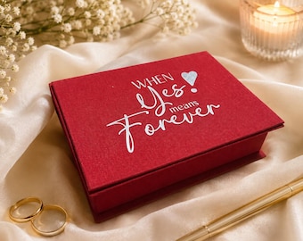 May include: A rectangular red box with a textured finish featuring the silver text "WHEN Yes! means Forever" and a small silver heart icon. The box is positioned on a draped cream fabric alongside two gold rings and a gold ballpoint pen.