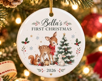 Könnte beinhalten: Ein runder, weißer Keramik-Weihnachtsbaumschmuck mit goldener Aufhängung. Das Motiv zeigt ein Rehkitz mit rotem Schal, ein Eichhörnchen, einen Hasen, einen Tannenbaum und Stechpalmen. Der Text lautet Bella's FIRST CHRISTMAS und 2026, verziert mit Sternen.