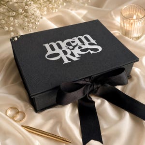 May include: A rectangular black box with a hinged lid featuring the white text "memories" printed on top. The box is tied with a wide black satin ribbon bow. Two gold-toned rings and a gold-toned pen are placed nearby on a soft white fabric.