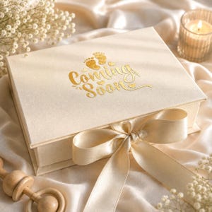 May include: A rectangular cream-colored gift box featuring the gold foil text Coming Soon and two small baby footprints. The box is tied with a wide matching satin ribbon bow on the front lid, creating a decorative closure for the container.