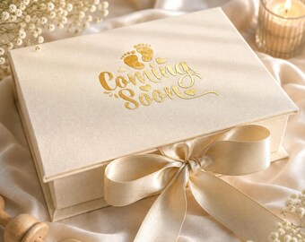 May include: A rectangular cream-colored gift box featuring the gold foil text Coming Soon and two small baby footprints. The box is tied with a wide matching satin ribbon bow on the front lid, creating a decorative closure for the container.