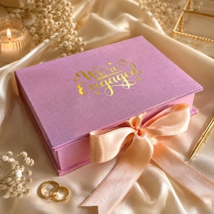 May include: A rectangular lavender gift box with a hinged lid featuring the gold foil text "We're Engaged" and a small ring icon. The box is tied with a wide, pale peach satin ribbon bow. Two gold metal rings are placed near the box.