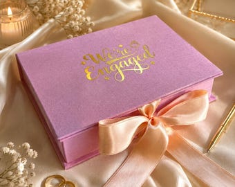 May include: A rectangular lavender gift box with a hinged lid featuring the gold foil text "We're Engaged" and a small ring icon. The box is tied with a wide, pale peach satin ribbon bow. Two gold metal rings are placed near the box.