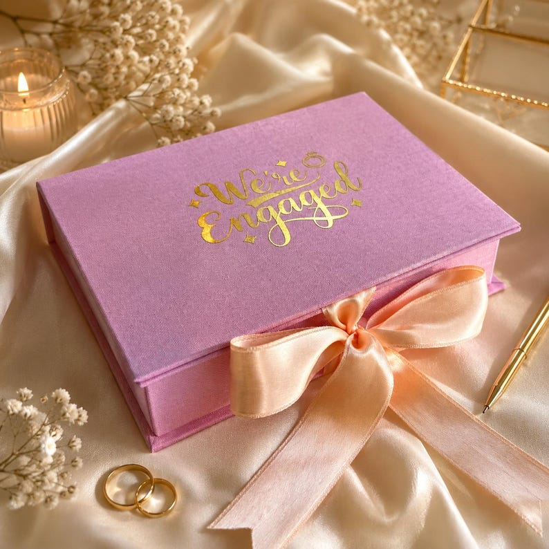 May include: A rectangular lavender gift box with a hinged lid featuring the gold foil text "We're Engaged" and a small ring icon. The box is tied with a wide, pale peach satin ribbon bow. Two gold metal rings are placed near the box.