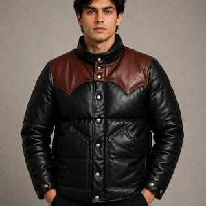 Può includere: Un piumino nero caratterizzato da uno sprone in pelle marrone sul petto e sulle spalle. La giacca presenta un colletto alto, una chiusura frontale con bottoni a pressione dorati, due tasche anteriori e maniche lunghe con polsini a pressione.