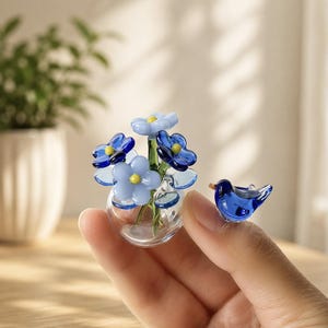 Op de afbeelding: Een miniatuur glazen sculptuur bestaande uit een heldere bolvormige vaas met diverse lichtblauwe en donkerblauwe bloemen met gele harten, vergezeld door een klein, massief donkerblauw vogelbeeldje naast de vaas.