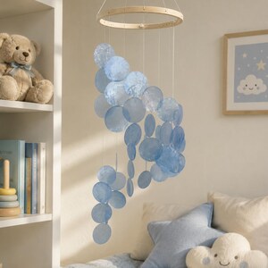 May include: A hanging mobile featuring a circular wooden frame with multiple translucent, light blue, circular discs suspended by thin white strings. The discs vary in size and are arranged in a cascading, spiral pattern, creating a layered effect.