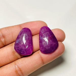 以下が含まれることがあります： 深いマゼンタパープルの色合いをした、ルースでファセットカットされた洋梨型のルビー2石。それぞれの宝石は、光を反射する複数の三角形のファセットを持つ平らで磨かれた表面が特徴で、半透明の質感と滑らかで丸みを帯びたベースを見せています。