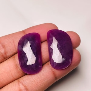 Pode incluir: Duas pedras preciosas semicirculares de cor púrpura profunda, facetadas e com acabamento polido. Cada pedra apresenta uma parte traseira plana e lisa e uma superfície frontal multifacetada que reflete a luz, exibindo uma cor violeta saturada.