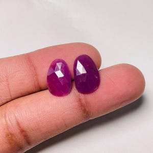 Op de afbeelding: Twee losse, ovaalvormige robijnen met een gefacetteerd oppervlak. De edelstenen hebben een diepe, verzadigde magenta-rode kleur en een gepolijste afwerking, met meerdere geometrische facetten die het licht reflecteren op hun oppervlak.