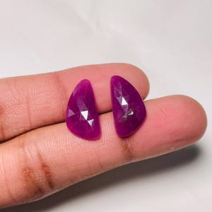 Op de afbeelding: Twee losse, gefacetteerde, halfronde edelstenen met een diepe magenta-roze kleur. Elke steen heeft een platte, gebogen rand en een puntige kant, met een geometrisch, veelzijdig oppervlak dat licht reflecteert over de gepolijste zijde.