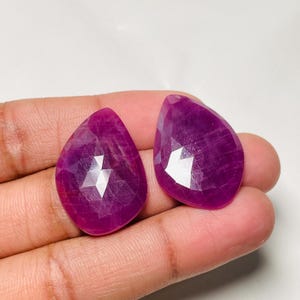 Puede incluir: Dos rubíes facetados en forma de pera con un tono magenta rosado intenso. Cada piedra preciosa presenta una superficie pulida con facetas geométricas que reflejan la luz, mostrando una calidad translúcida y un perfil de color vibrante y saturado.