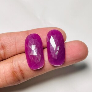 Pode incluir: Duas pedras preciosas de cor magenta, de formato oval e facetadas com uma superfície polida. As pedras apresentam um corte geométrico que reflete a luz nas suas superfícies translúcidas de cor rosa-púrpura profundo, com um perfil de borda arredondado.