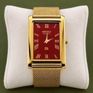 Könnte beinhalten: Eine rechteckige, goldfarbene Seiko Quartz Armbanduhr mit einem tiefroten Zifferblatt und goldenen römischen Ziffern als Stundenmarkierungen. Die Uhr hat ein goldenes Mesh-Armband aus Metall und eine Krone an der rechten Seite.