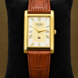 Op de afbeelding: Een rechthoekig Seiko Quartz horloge met een goudkleurige kast en een bruine lederen band met krokodillenmotief. De witte wijzerplaat is voorzien van zwarte Romeinse cijfers, gouden wijzers en een centraal gestructureerd patroon.