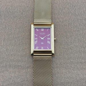 Könnte beinhalten: Eine rechteckige Seiko Quartz Armbanduhr mit einem leuchtend violetten Zifferblatt, silberfarbenen römischen Ziffern als Stundenmarkierungen und Zeigern. Die Uhr hat ein goldfarbenes Metallgehäuse und ein passendes feines Gliederarmband.