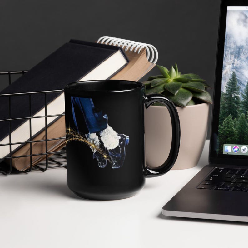 K&ouml;nnte beinhalten: Schwarze Kaffeetasse mit einer Grafik eines Fu&szlig;es in einem schwarzen Schuh, einer wei&szlig;en Socke und einer blauen Hose, mit einer goldenen Glitzerspur. Die Tasse hat einen schwarzen Griff und ein gl&auml;nzendes Finish.