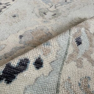 May include: A woven area rug featuring a light blue, beige, and charcoal gray geometric pattern. The rug is folded over to reveal the textured, knotted pile construction and the underside of the fabric, showcasing a repeating medallion design.