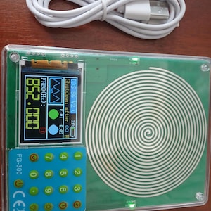 Puede incluir: Un generador de señales FG-300 verde con carcasa protectora transparente, pantalla digital que muestra 852.000 FREQ (Hz), teclado azul con números del 0 al 9 y un gran patrón de bobina en espiral blanca. Se incluye un cable de carga USB blanco.