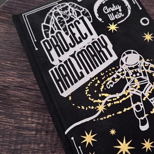 May include: A black book with the title "Project Hail Mary" in white, featuring an astronaut illustration and star accents. The author's name, Andy Weir, is also visible. The book is set against a dark wooden surface.